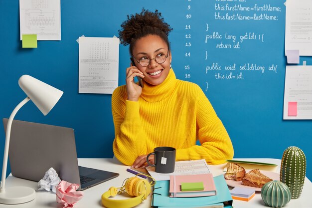 Positive dark skinned girl makes telephone call, discusses improvement and developing business project, dressed in yellow sweater, looks aside, poses against blue background