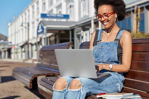 Positive dark skinned girl traveler blogger enjoys online communication, makes video call, talks with friend from abroad, uses laptop, earphones, sits at bench near railway station waits for transport