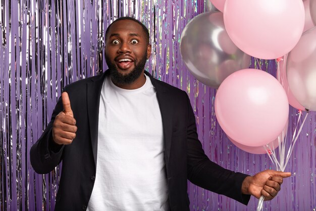 Positive dark skinned guy gives thump up, likes brilliant idea of organizing party, holds bunch of air balloons, has widely opened eyes, celebrates special occasion