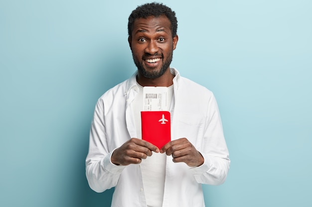 Free photo positive dark skinned man with thick bristle, holds fly ticket and passport, prepares for tour abroad, waits for plane departure, wears white fashionable clothes, models over blue wall