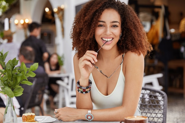Free photo positive dark skinned mixed raced female with curly bushy hairstyle holds eyewear in hands, wears casual t shirt, has lunch or coffee break in cafe