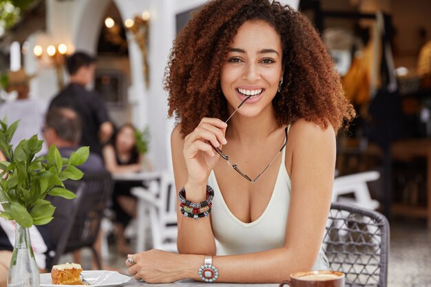 Positive dark skinned mixed raced female with curly bushy hairstyle holds eyewear in hands, wears casual t shirt, has lunch or coffee break in cafe
