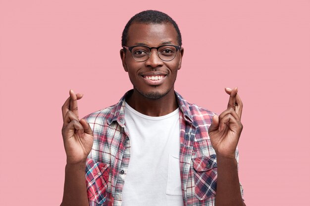 Positive dark skinned student wonk crosses fingers before passing exam, dressed in casual checkered shirt, has happy expression