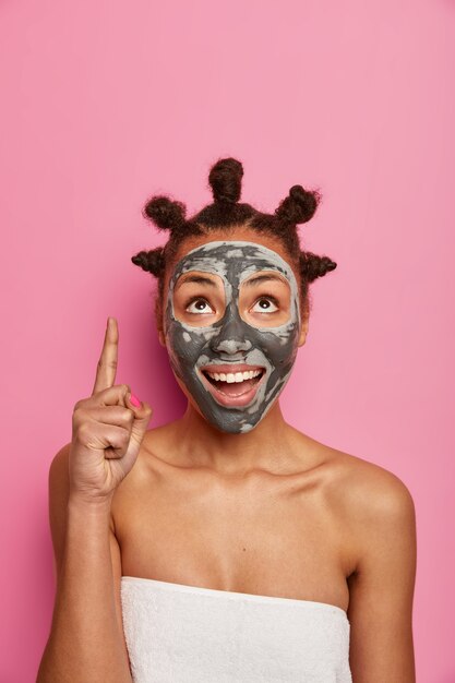 Positive dark skinned woman indicates upwards, shows blank space for advertisement, does beauty treatments, applies clay mask, has bare shoulders, wears soft towel around body. Wellbeing concept