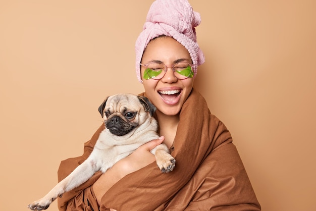 Positive dark skinned woman laughs happily wrapped in blanket poses with pug dog undergoes beauty procedures wears spectacles applies green hydrogel patches isolated over beige wall Domestic animals