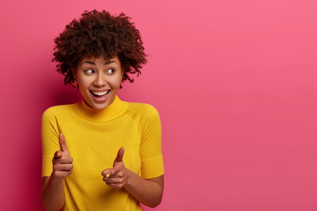 Positive dark skinned young female points , chooses you, feels happy and relaxed, giggles gladfully, looks aside, wears yellow t shirt, poses against pink  wall, copy space aside