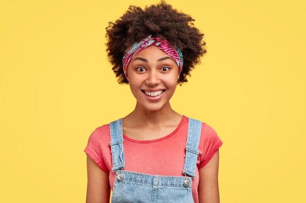 巻き毛のふさふさした髪のポジティブなダークスキンの若い女性は、歯を見せる笑顔が広く、心地よいものを聞いて喜んでおり、デニムのオーバーオールを着て、黄色い壁に立っています。感情の概念