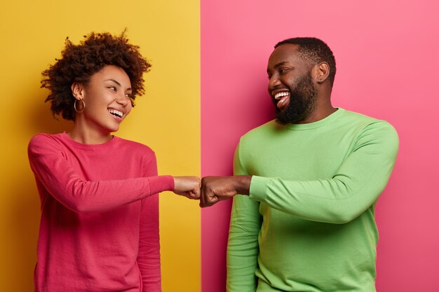 Позитивные темнокожие молодая женщина и мужчина бьют кулаками, соглашаются быть одной командой, счастливо смотрят друг на друга, празднуют выполненное задание, носят розовую и зеленую одежду, позируют в помещении, заключают успешную сделку