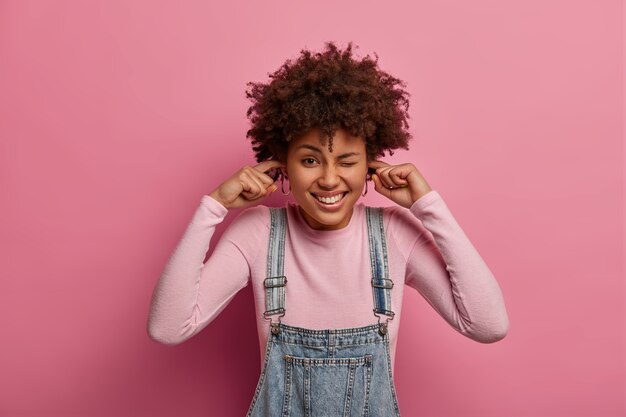 Free photo positive dark skinned young woman winks eyes and plugs ears, unwilling to listen loud noise, comes on noisy party, being in good mood, pretends not to hear, isolated over rosy pastel wall