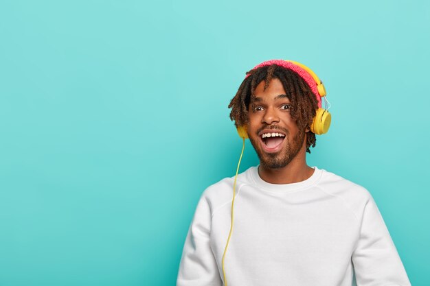 Positive dark skinned youngster has dreadlocks,  wears pink knitted hat and white jumper, models against blue wall, copy space for text