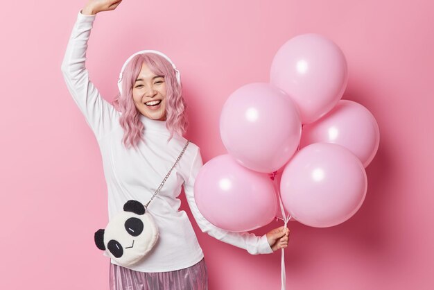 Positive delighted Asian woman with rosy hair lost in music dances happily holds bunch of helium balloons expresses happy emotions poses against pink background People fun and celebration concept