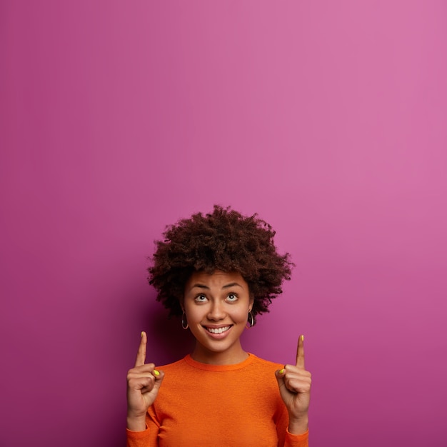 Free photo positive delighted curly woman gets new idea for development, points index fingers above, shows copy space on purple wall, smiles pleasantly, poses indoor, demonstrates nice offer for you