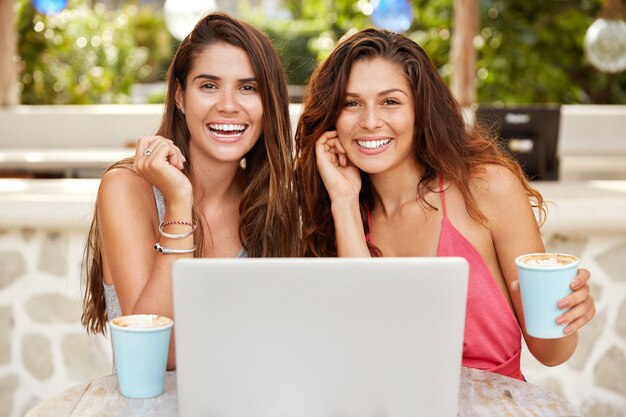 Positive delighted females with pleasant appearance, watch movie on laptop computer, drink fresh coffee, sit at cozy terrace cafe, have happy expressions.