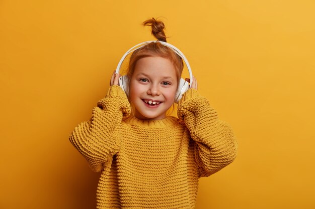 Positive delighted ginger girl listens audio track via headphones, enjoys favorite hobby, dressed in oversized knitted sweater, isolated over yellow wall. Children, music and fun concept