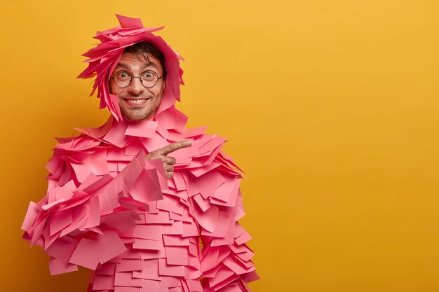 Positive delighted man points at product, advertises items for office, glad about advertisement, wears round spectacles, sticky notes over body and head, has happy smile, isolated on yellow wall