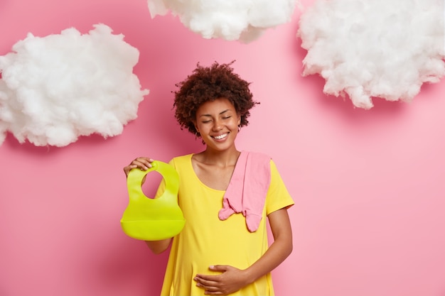 ポジティブな喜びの女性は、生まれたばかりの子供を期待し、おなかを注意深く抱きしめ、よだれかけと赤ちゃんの服を持って、彼女の妊娠を思い出すために写真撮影をします。期待と母性の概念