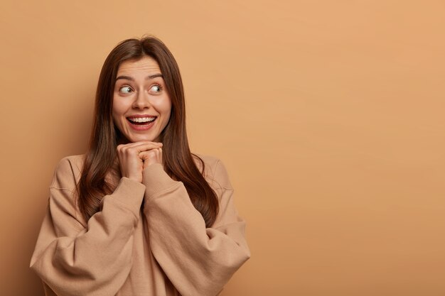 Positive delighted woman looks gladfully aside, has tender smile, keeps both hands under chin, has appealing appearance, wears sweatshirt