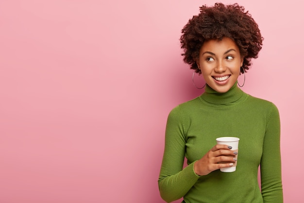 Positive dreamy female model poses with takeout coffee, warms up with hot drink, looks on left side, has casual talk
