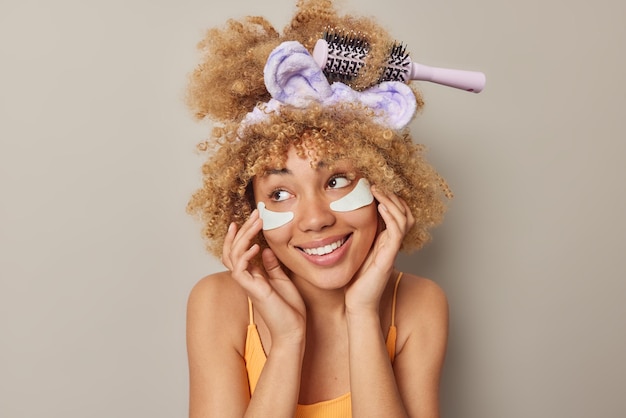 Foto gratuita la giovane donna sognante positiva con i capelli ricci folti applica cerotti di bellezza sotto gli occhi per ridurre i problemi di occhiaie e rughe indossa la fascia isolata su sfondo grigio concetto di cura della pelle