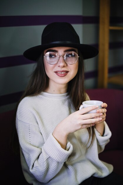 Positive elegant young woman in hat and eyeglasses with mug of drink