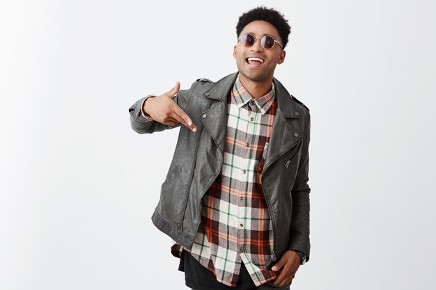 Positive emotions. Beautiful young joyful dark-skinned african guy with curly hair in fashionable black leather jacket and tan-glasses smiling with teeth, gesticulating with hand after seeing old frie