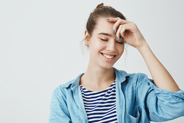 Positive emotions concept. Attractive woman with bun hairstyle holding hand on forehead while smiling with closed eyes, standing . Young artist creating new painting in mind