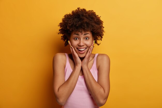 긍정적 인 감정 개념. 기쁜 즐거운 곱슬 아프리카 계 미국인 여자는 뺨을 만지고, 예기치 않은 멋진 것을 배웠고, 행복한 미소로 보이며, 노란색 벽 위에 포즈를 취합니다.