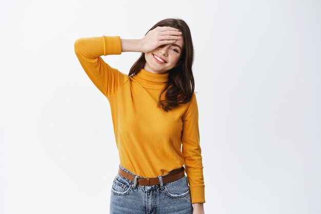 Positive emotions Happy and carefree young woman holding hand on face smiling and looking cheerful at camera standing on white background