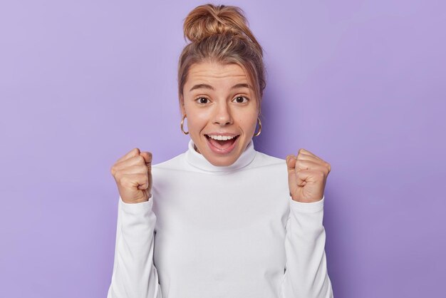 Positive emotions and human reactions concept. Young happy woman cleches fists exclaims loudly encourages someone or supports favorite team wears white turtleneck isolated over purple studio wall