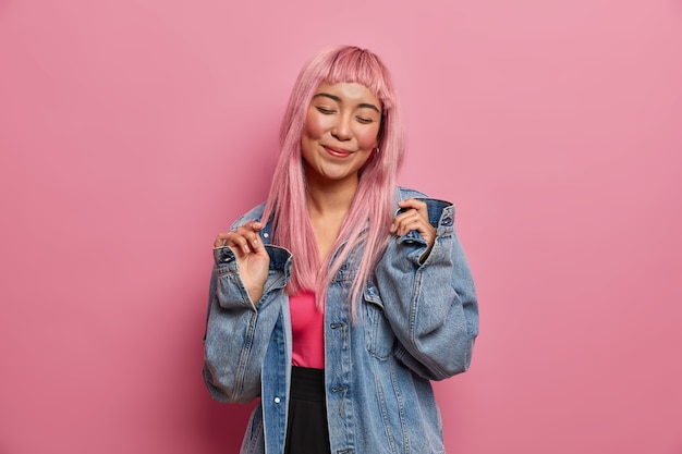 Positive emotions, people and fashion concept. Smiling glad young woman with pink long hair, closes eyes, happy to buy new denim jacket, stands indoor