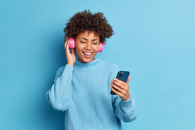 Positive entertained millennial girl with curly hair holds smartphone makes video call wears stereo headphones for listening favorite music dressed in warm jumper 