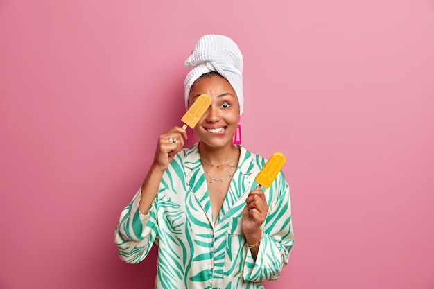 Positive ethnic housewife with dark skin covers eye with tasty cold ice cream has fun while eating delicious sweet dessert wears domestic clothes wrapped bath towel on head. Summer time concept