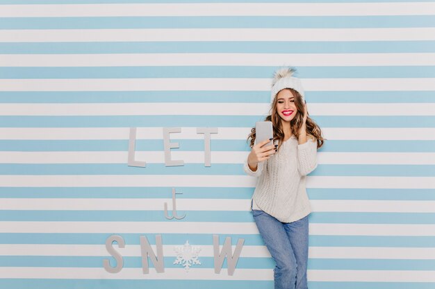 Positive European female model with pink lipstick sincerely smiles and happily makes selfies near striped wall