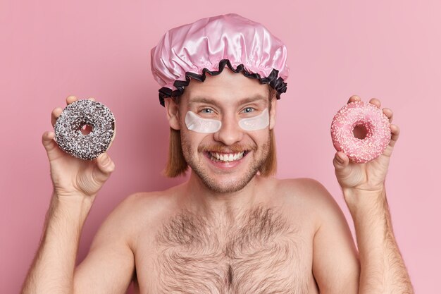 Позитивный европейский парень с радостным выражением лица накладывает коллагеновые пятна под глаза, держит сладкие пончики, носит шапочку для душа, стоит полуголым