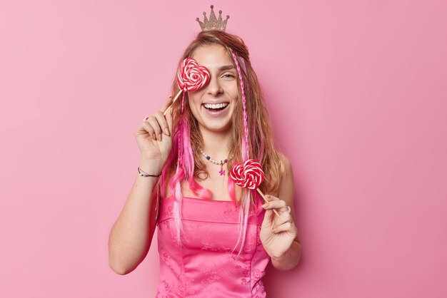 Foto gratuita la donna europea positiva copre l'occhio con lecca-lecca a forma di cuore indossa un abito festivo e una piccola corona sulla testa sorride ampiamente ha denti bianchi perfetti posa su sfondo rosa tempo per la festa