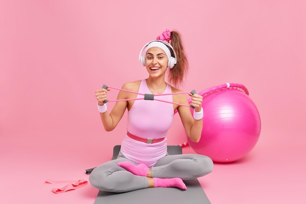 スポーツ服を着たポジティブなヨーロッパの女性は、抵抗バンドを伸ばし、フィットネスマットの上に足を組んで座ります。運動器具を使用して、ピンクの壁にヘッドホンでポーズをとって音楽を聴きます。