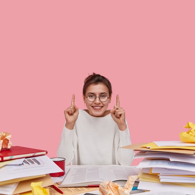 Positive European woman with glad facial expression, dressed in white casual clothes, shows direction upside, uses paper documents, has toothy smile