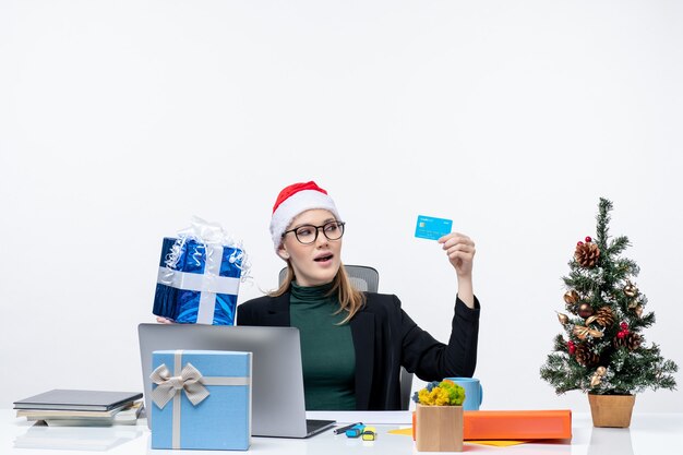 サンタクロースの帽子と白い背景の上のクリスマスの贈り物と銀行カードを保持しているテーブルに座って眼鏡をかけてポジティブ興奮ビジネス女性