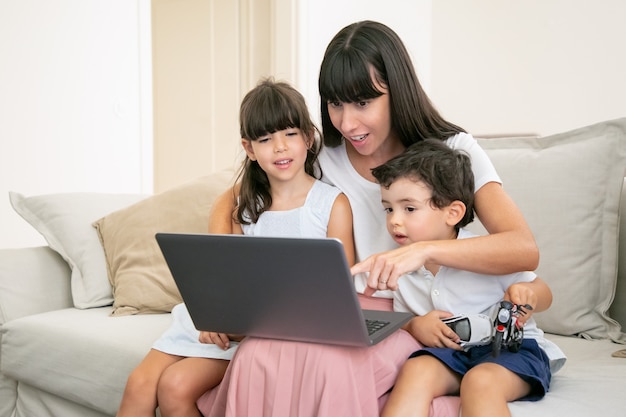 Positive excited mother embracing two kids and pointing at laptop display. Family sitting on couch at home and watching movie.