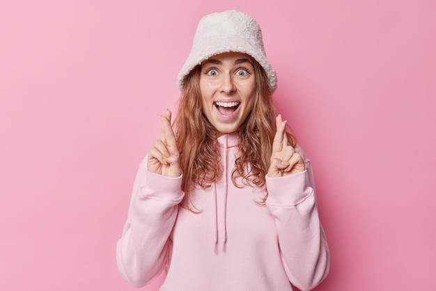Positive excited woman keeps fingers crossed wishes dreams come true waits for news laughs happily wears panama and casual hoodie isolated over pink background Hopefull cheerful millenial girl