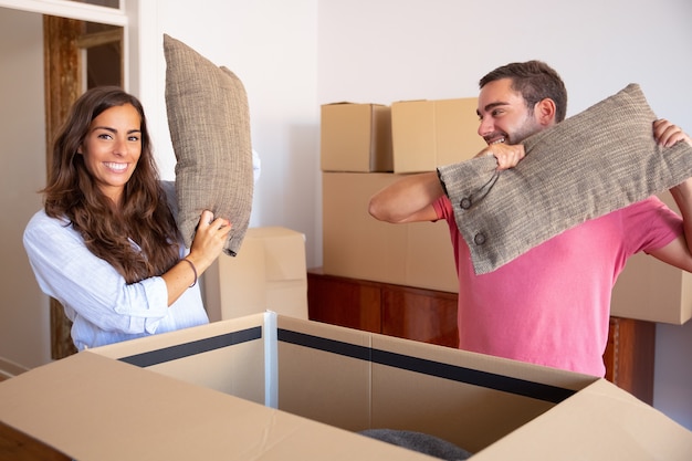 Позитивно возбужденные молодой мужчина и женщина достают подушки из открытой картонной коробки, наслаждаются перемещением и распаковкой вещей