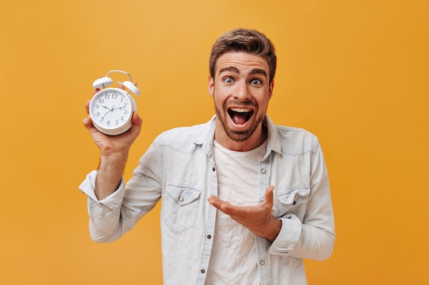 Positive fashionable guy with brown hair and red beard in white tshirt and denim modern jacket showing at big clock and looking into camera