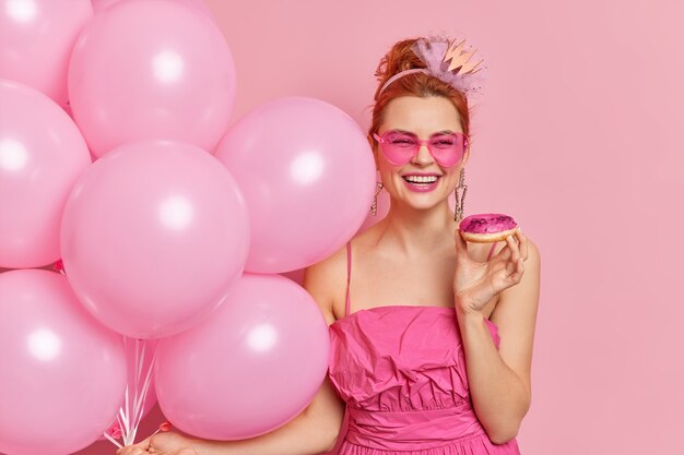 Позитивная модная рыжая женщина широко улыбается, имеет праздничное настроение, держит надутые воздушные шары из вкусных пончиков, носит солнцезащитные очки в форме сердца и платье
