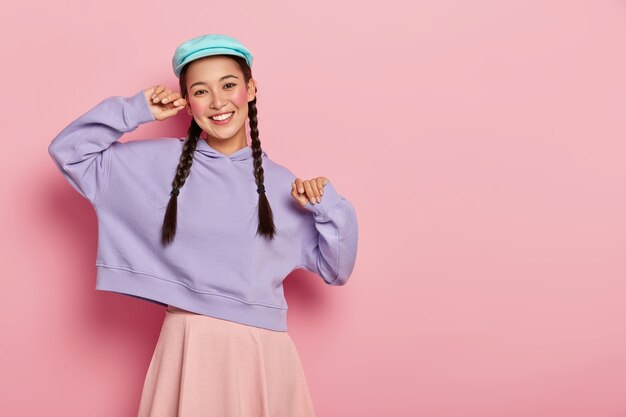 Бесплатное фото Позитивная модная девочка-подросток с макияжем, в стильной кепке, фиолетовой толстовке и юбке, в хорошем настроении, приходит на встречу с однокурсниками, изолирована за розовой стеной