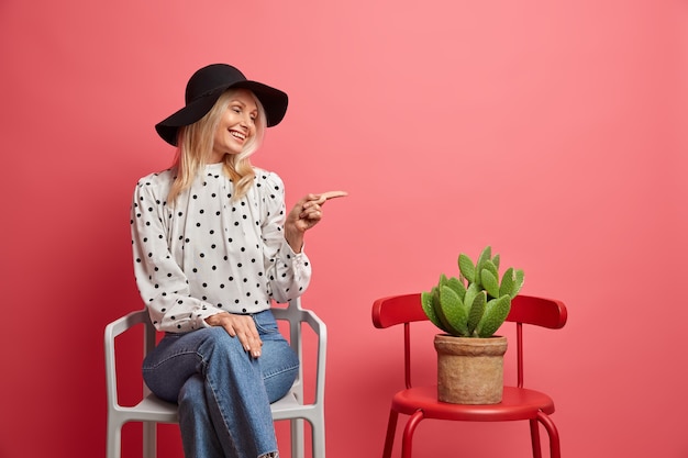 Positive fashionable woman points at potted cactus poses on chair indoor