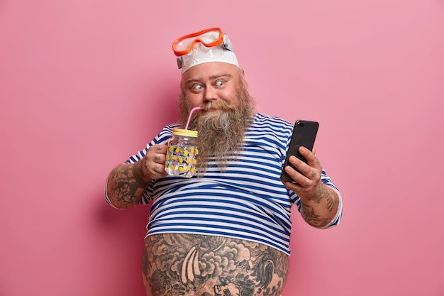 Positive fatso guy drinks fresh water, takes photo via cellphone, wears swimming snorkeling mask, undersized sailor shirt