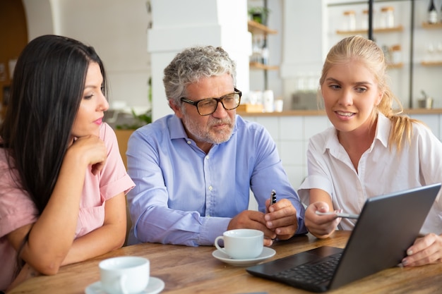 Positive female agent showing project presentation on laptop to young woman and mature man, pointing pen at display, explaining details