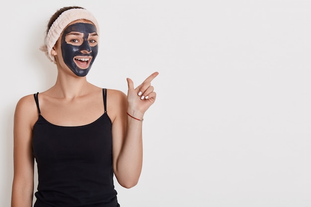 Positive female applies nourishing mask on face, pointing fore finger aside on copy space, undergoes beauty treatments, poses indoor against white wall. Copy space.