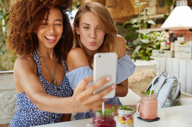 陽気な女性の親友が自分撮りのポーズをとる、現代の携帯電話を使用する、レストランに座る、面白い表現をする、ソーシャルネットワークで写真を共有する、ワイヤレスインターネットに接続する。友情の概念
