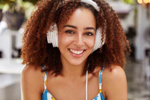 Positive female blogger with dark skin and Afro hairstyle recreats outdoor during listening to favourite music, connected to cell phone